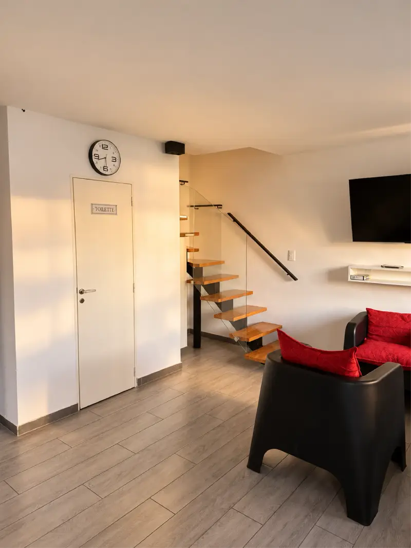 Living del dúplex con piso de madera clara, escalera de peldaños de madera con baranda de vidrio, sillones negros con almohadones rojos, televisor y la puerta del toilette.