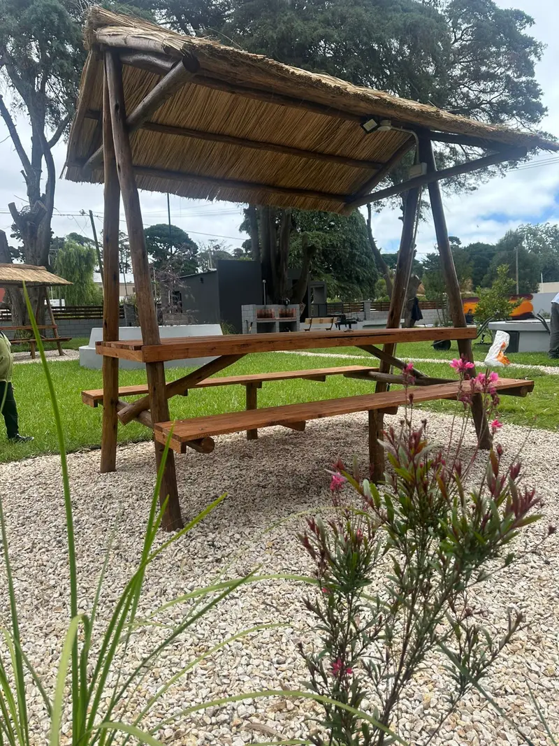 Mesa de madera con bancos bajo un techo de paja, sobre piedra partida, con el parque y árboles altos alrededor.