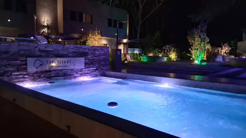 La pileta de noche, iluminada, con plantas y luces cálidas.